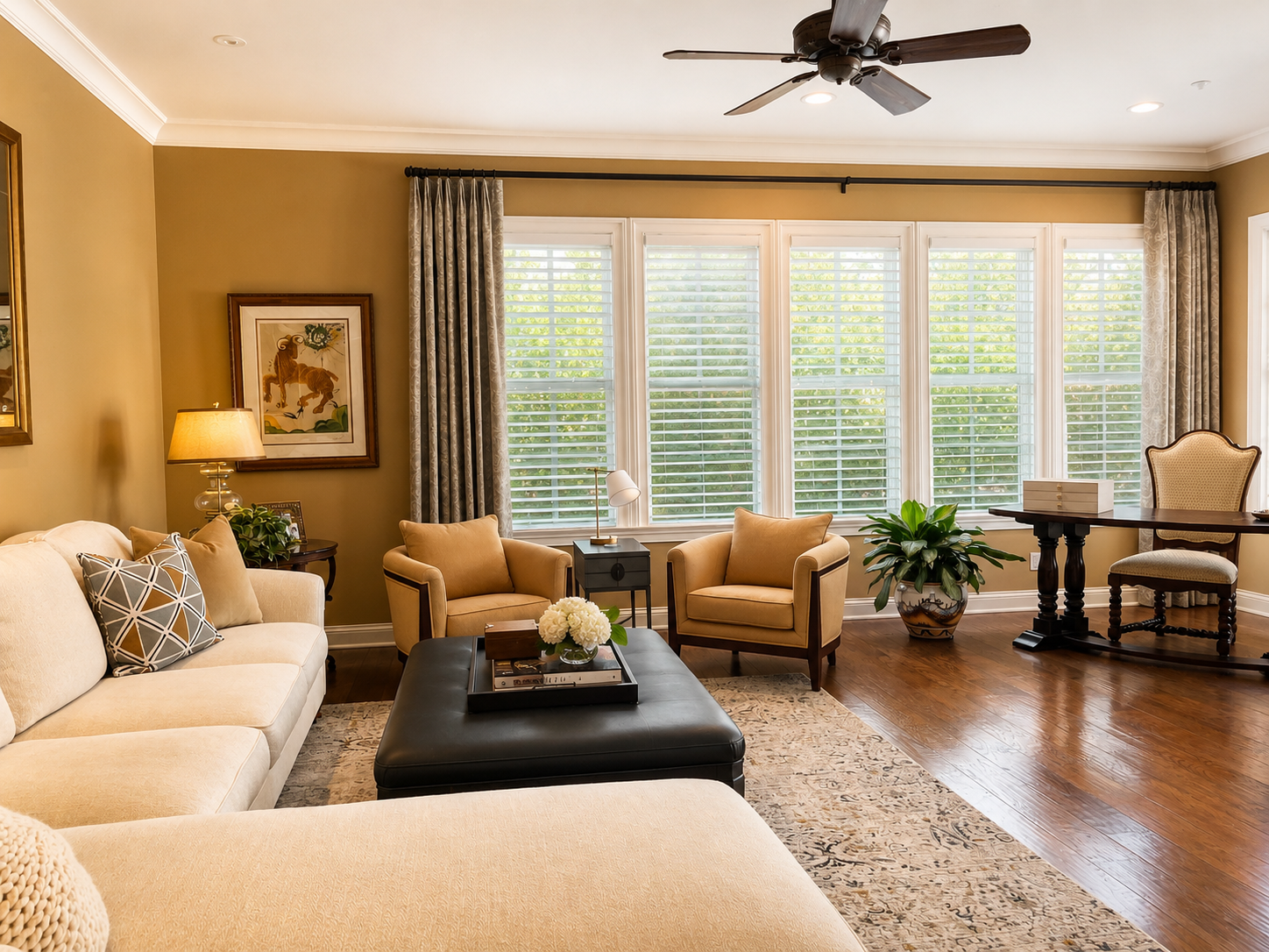 A living room with beige walls and large windows with white blinds, decorated with framed artwork and several green plants. The room features a beige sofa with patterned pillows, two matching armchairs, a black ottoman with a tray and flowers, and a 