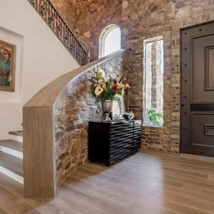 Interior entryway with exposed brick walls, a staircase with a beige marble handrail, a black console table with a large floral arrangement, and an ornate dark front door.