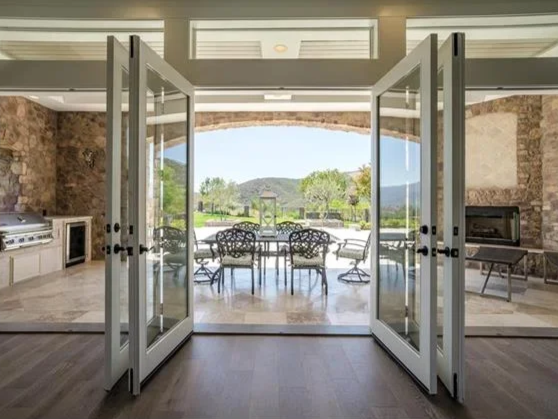 Open glass doors leading to an outdoor patio with a table and chairs, overlooking a green yard and distant hills.