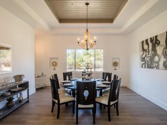 A dining room with a round table and eight chairs, a chandelier hanging above, a window with view of trees, and wall decorations including a large abstract painting.