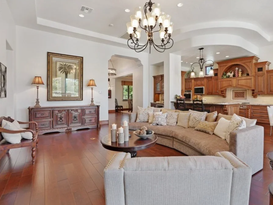 Interior of a living room with wooden flooring, a cream-colored sectional sofa with multiple cushions, a round wooden coffee table with candles, a wooden sideboard with lamps, and a kitchen area in the background with wooden cabinets and pendant ligh