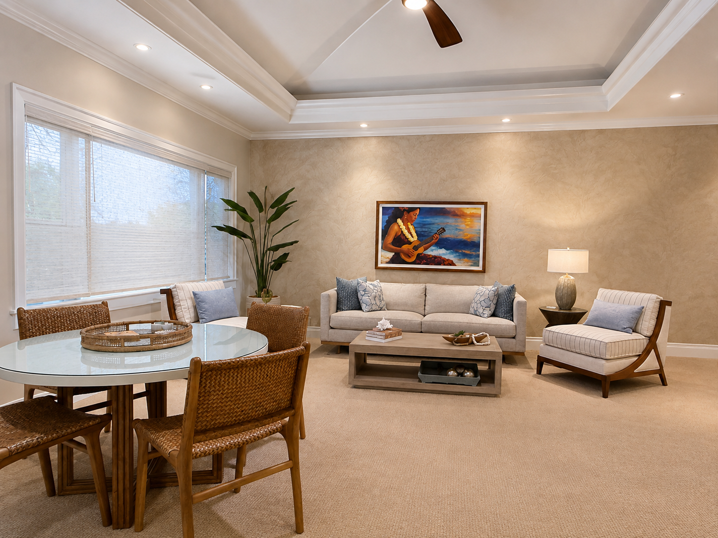 A living room with a dining area, featuring a white round table with four wicker chairs, a beige sofa with pillows, a chair with a pillow, a floor lamp, a painting of a woman playing a ukulele, a large window with blinds, a tall plant, and a ceiling fan.