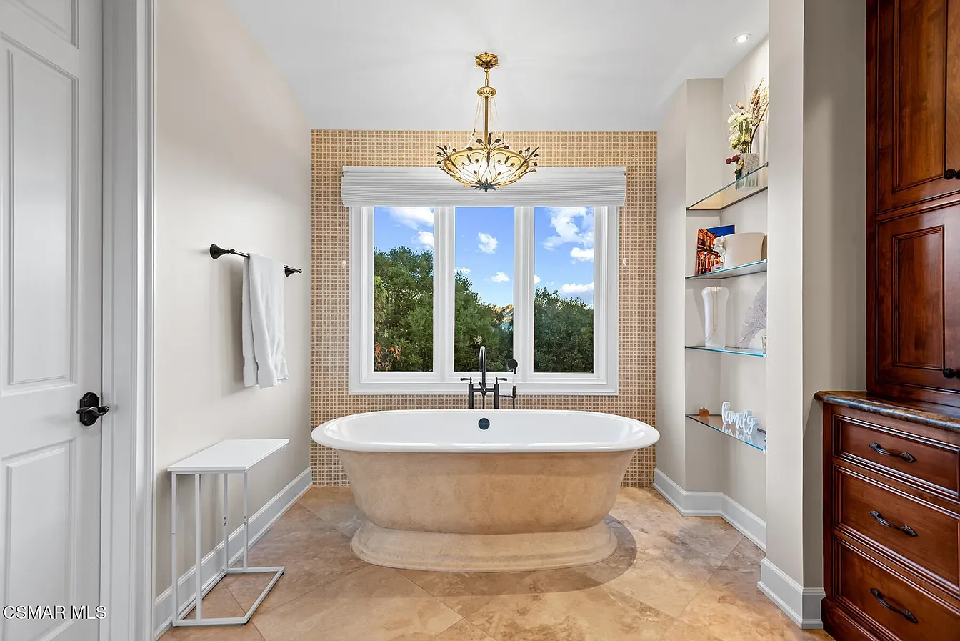Bathroom with a free-standing bathtub in front of a large window showing trees and blue sky. The room has a chandelier, a white towel on a black towel bar, and built-in shelves with decorative items. The floor is tan tile, and there is a tall wooden 
