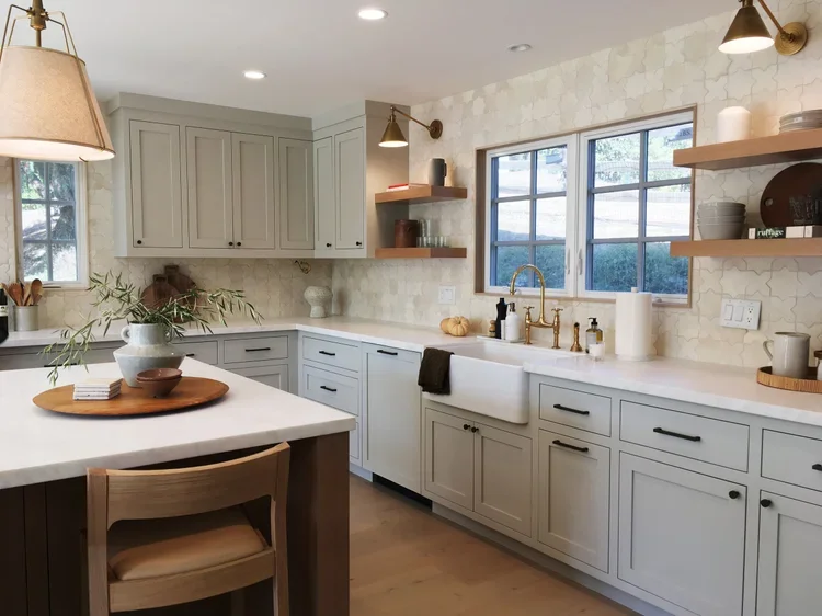 A modern kitchen with light gray cabinets, white countertops, a farmhouse sink, and open wooden shelves with dishes and decor. There is a window above the sink, and a large kitchen island with a wooden tray and bowl on top. The room is well-lit with 