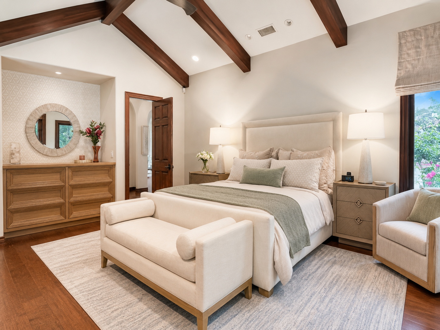 A cozy bedroom with a large bed, beige upholstered headboard, and matching nightstands with lamps. A light green throw blanket and pillows decorate the bed. There's a cream-colored bench at the foot of the bed, and a beige armchair with a green pillo