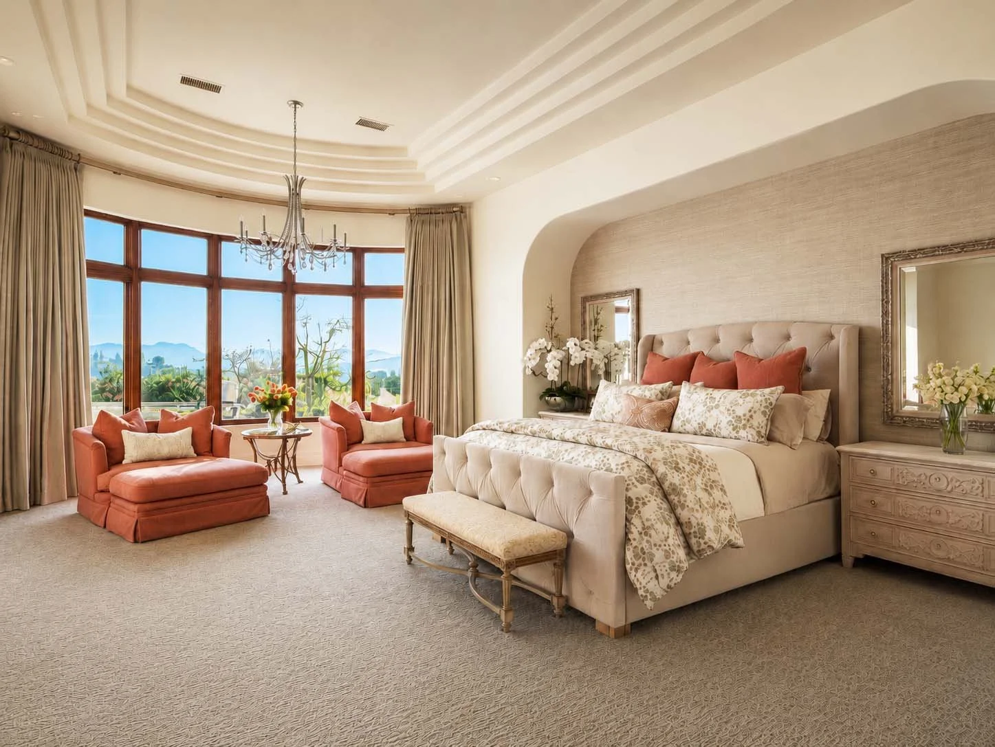 A spacious, elegant bedroom with a large bed, pink and white bedding, and matching pink armchairs. The room features a large window with a scenic view of trees and mountains, beige curtains, a chandelier, and floral decorations.