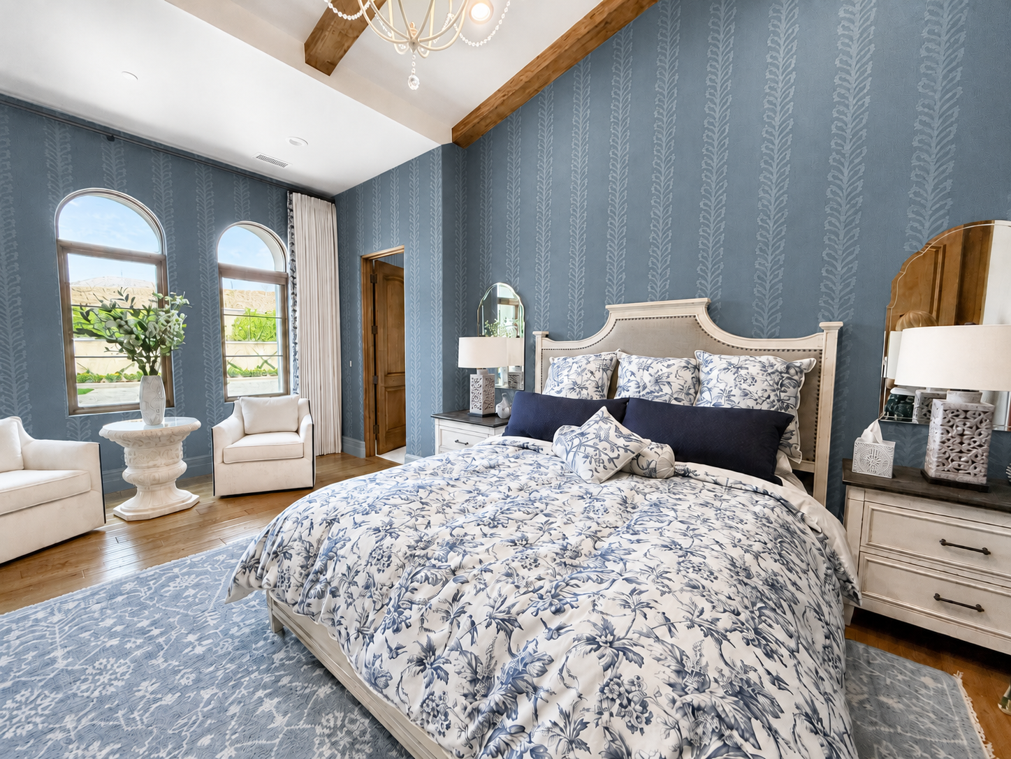A bedroom with blue patterned wallpaper, a large bed with floral bedding, multiple pillows, wooden furniture, and two arched windows with white curtains allowing outdoor views.