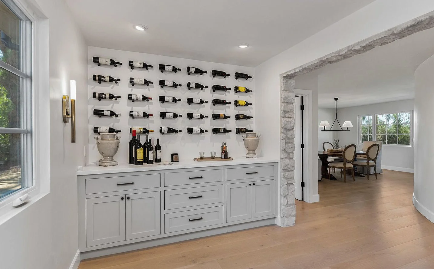 A wine storage and bar area with white cabinetry, wall-mounted wine racks holding bottles, and a small collection of bottles and glasses on the counter, with a stone archway leading to a dining room with large windows and a chandelier.