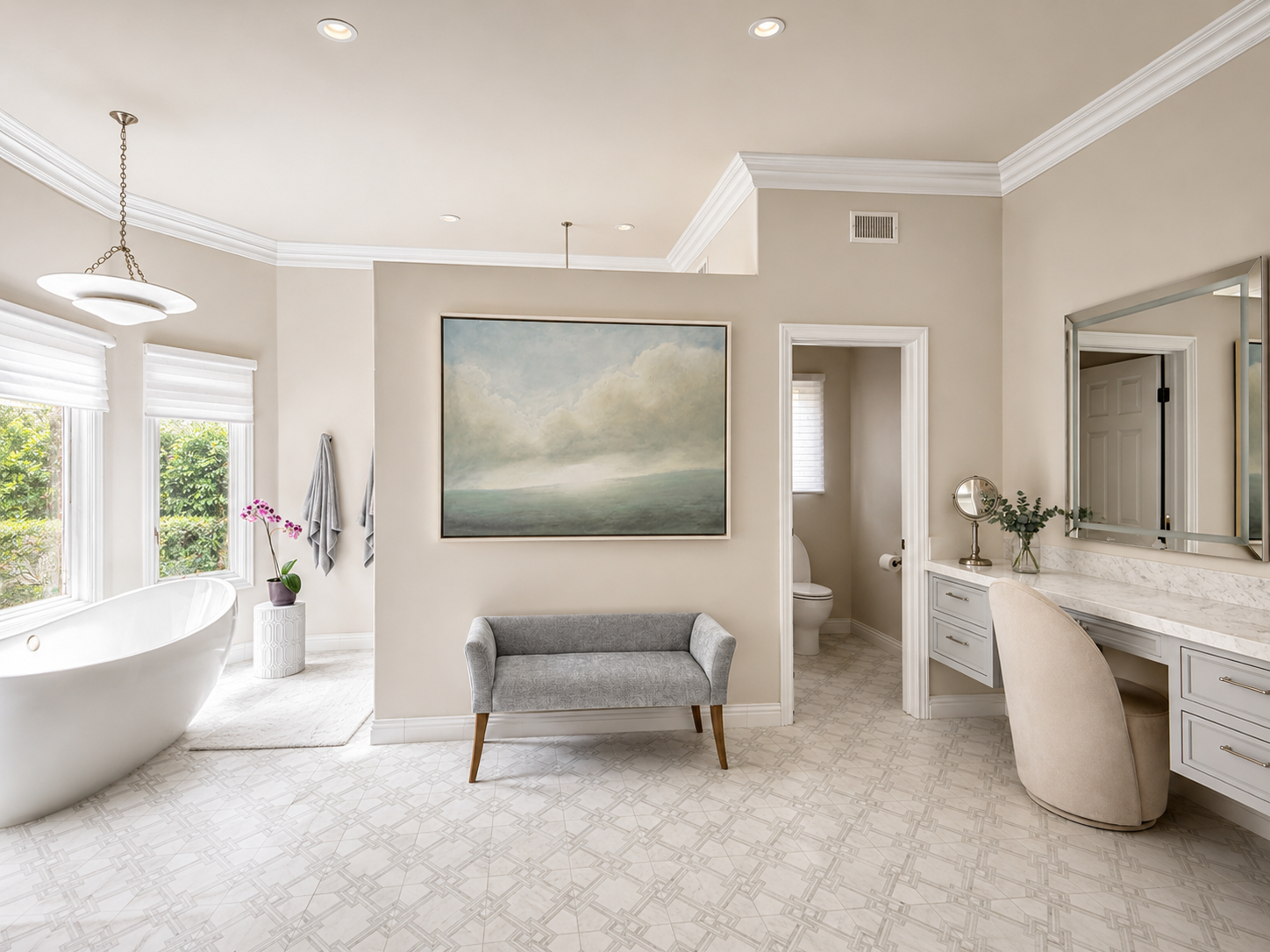 Bright bathroom with a freestanding bathtub near windows, a gray upholstered bench, artwork on the wall, a vanity with a mirror, and a toilet in a separate room.