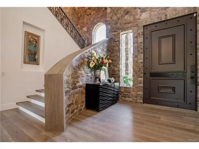 Interior entryway with exposed brick walls, a staircase with a beige marble handrail, a black console table with a large floral arrangement, and an ornate dark front door.