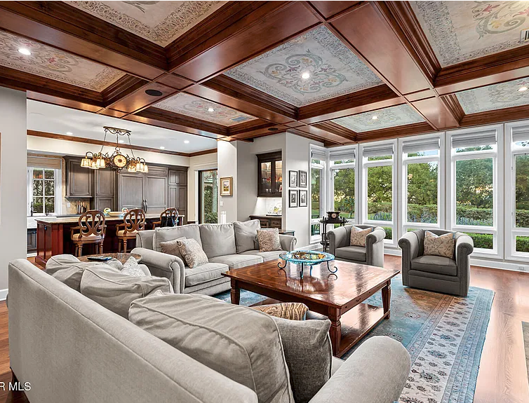 Spacious living room with large windows, gray sofas, a wooden coffee table, and a kitchen area with a dining table and chandelier.