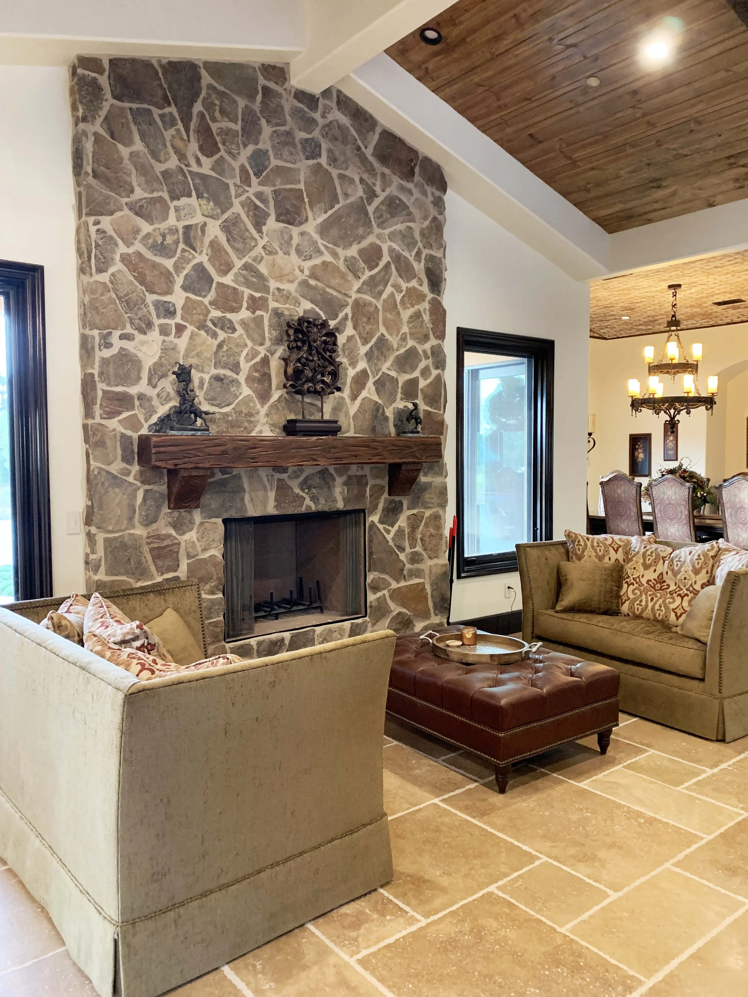 Living room with beige couch, floral patterned pillows, brown leather ottoman, stone fireplace with wooden mantel and decorative items, large window, and adjacent dining area with chandelier and upholstered chairs.