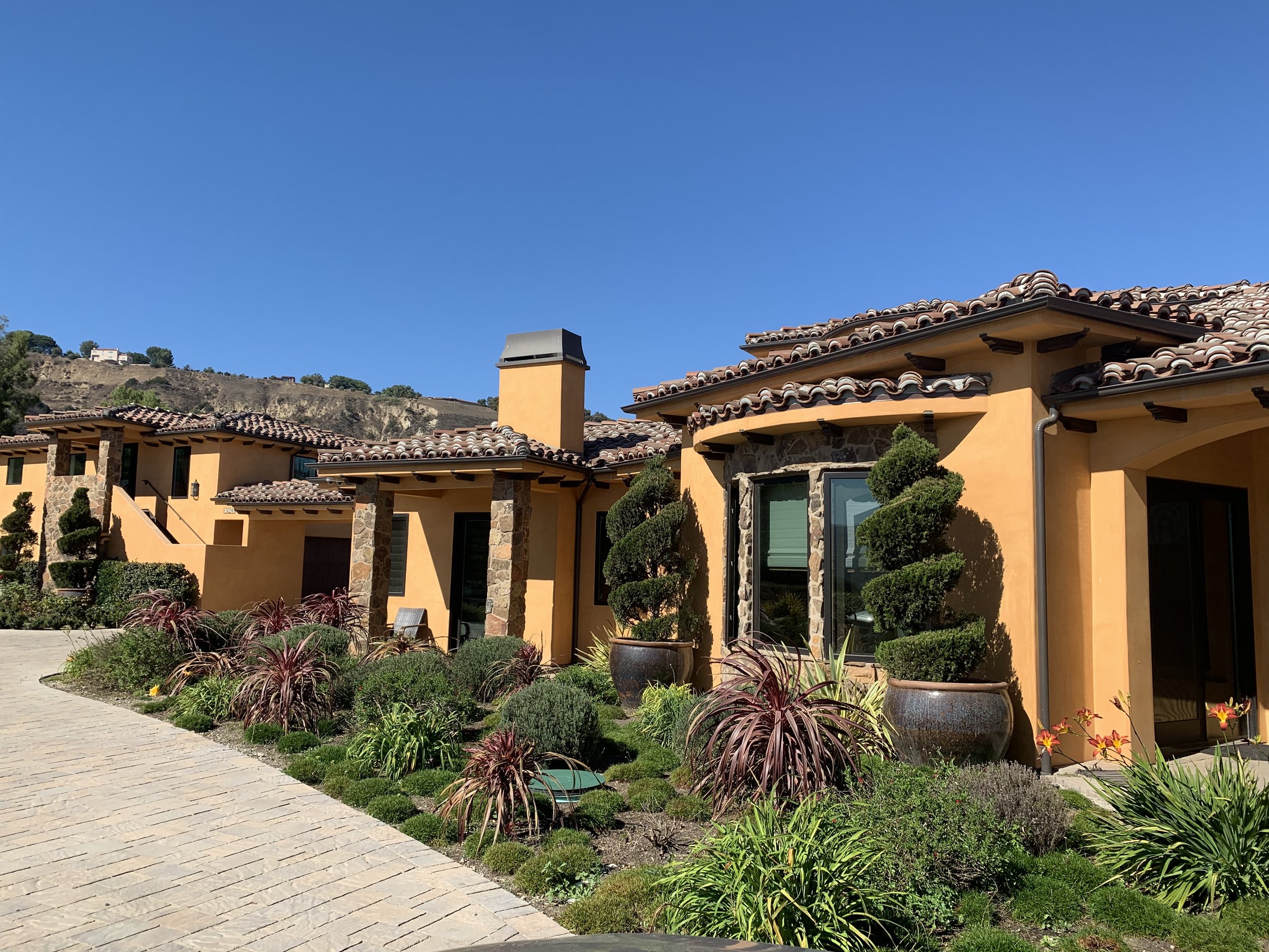 A view of a Mediterranean-style house with an orange stucco exterior, tiled roof, and lush landscaping in the foreground, with a hillside and clear blue sky in the background.