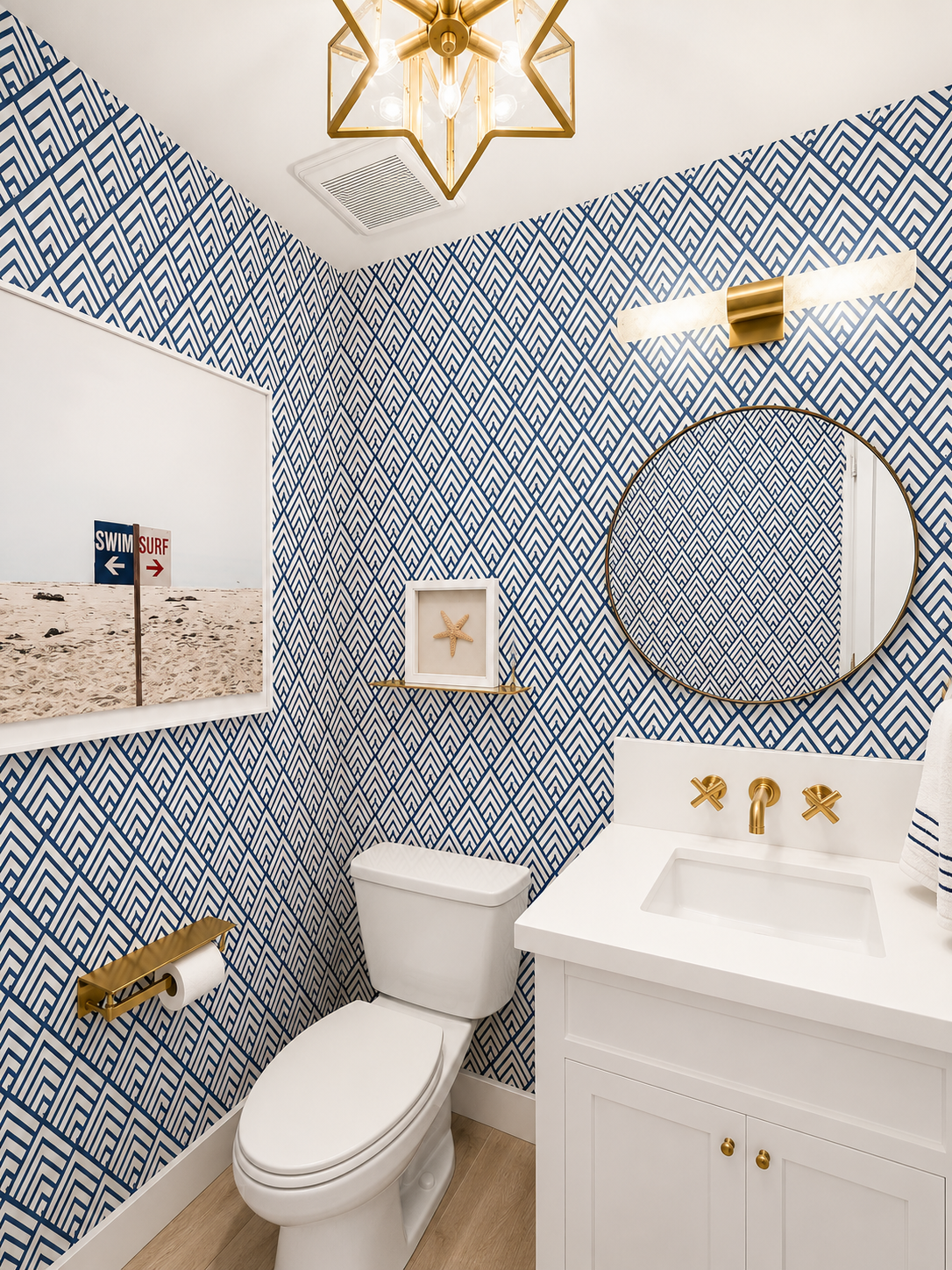 Bathroom with blue and white geometric patterned wallpaper, white toilet, white vanity with gold fixtures, large round mirror, wall art of a starfish, and a gold light fixture on the ceiling.