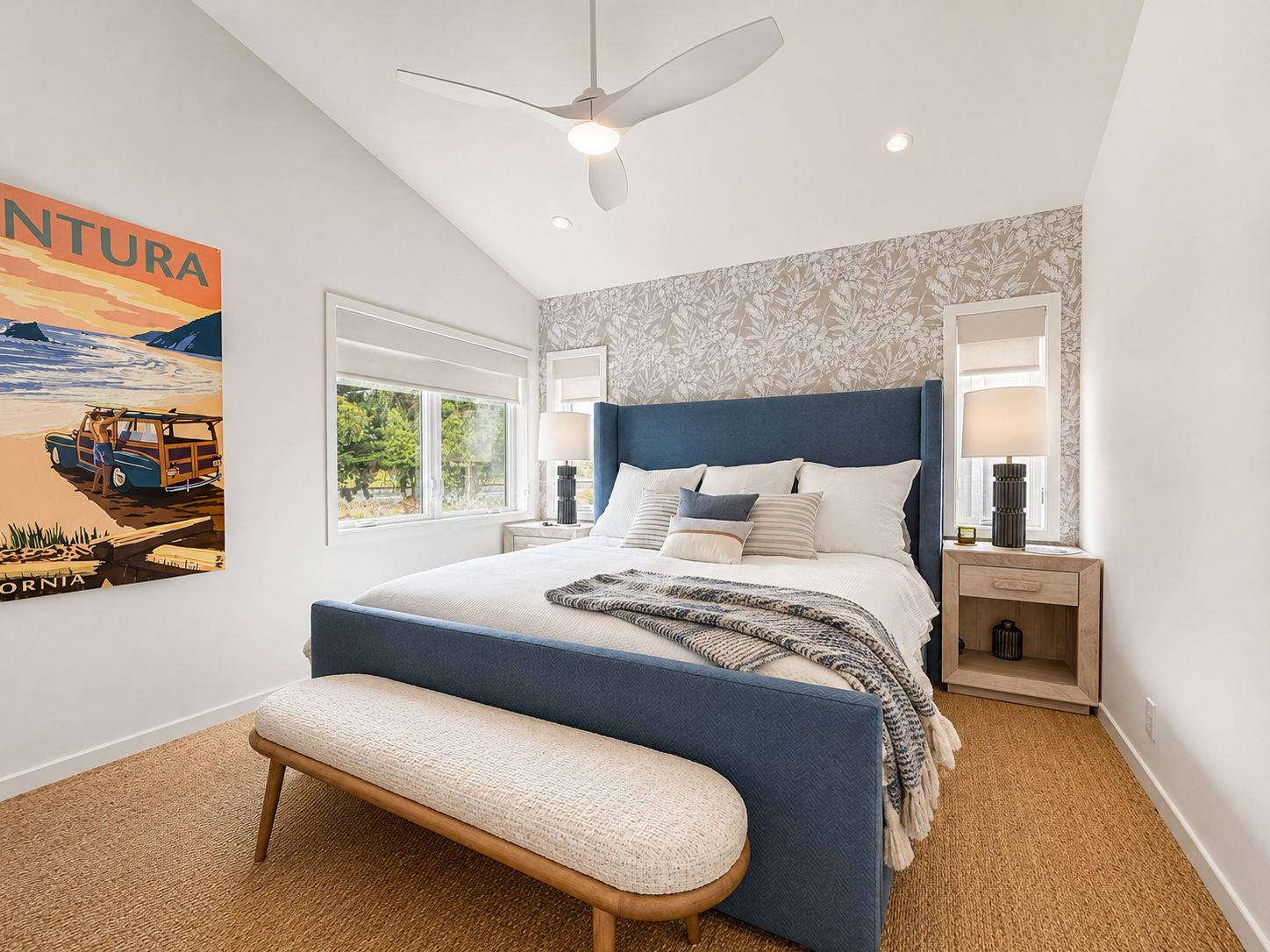 A bedroom with a blue upholstered bed, beige patterned wallpaper behind it, two nightstands with lamps, a large window, a ceiling fan, and a bench at the foot of the bed.