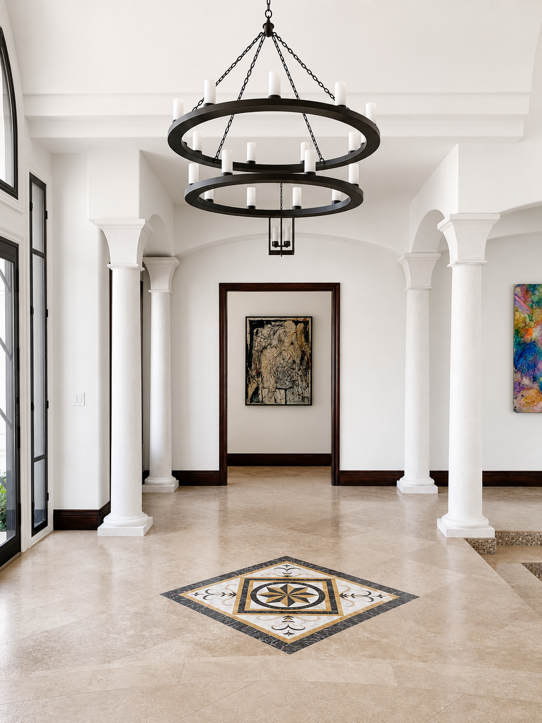 Interior view of a spacious, modern foyer with beige tiled floor featuring a decorative inlay, white pillars, and a black circular chandelier with candles hanging from the ceiling. Artwork is displayed on the walls.