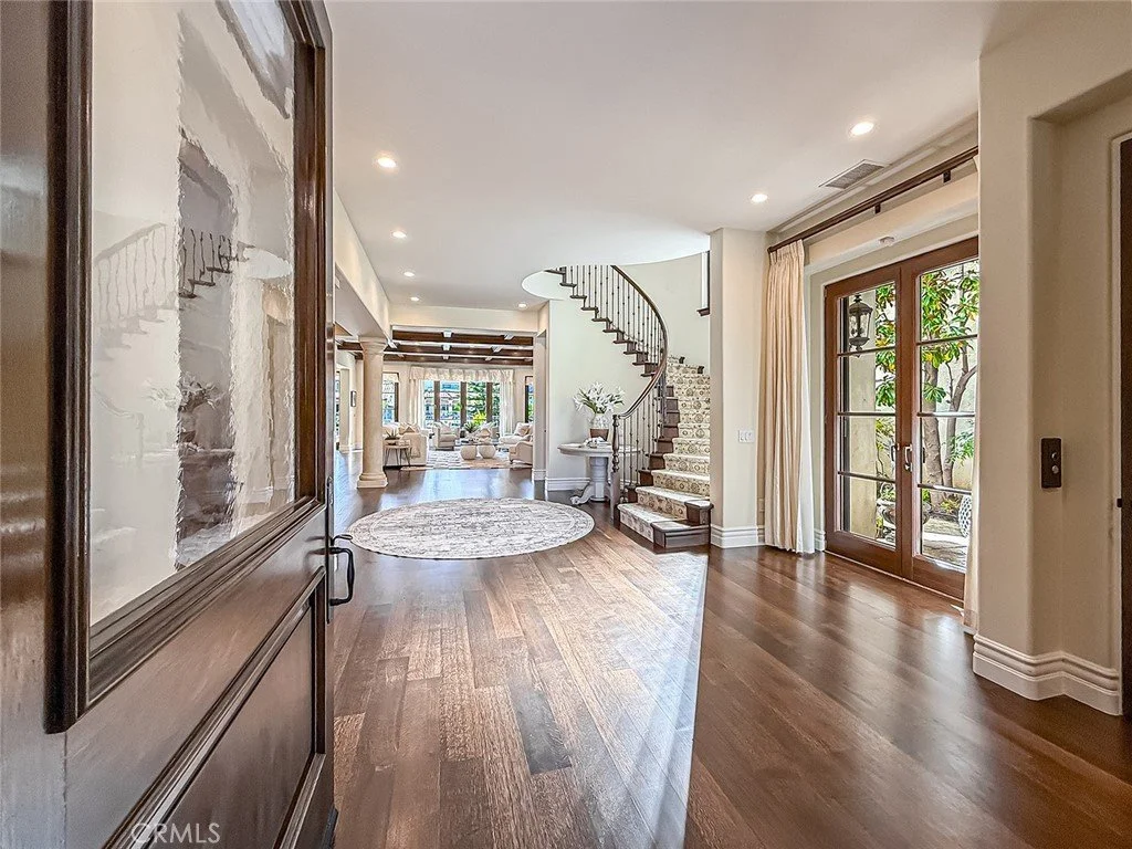 View of a spacious, well-lit living area with hardwood floors, a round area rug, a staircase with a curved handrail, and large glass doors leading outside.
