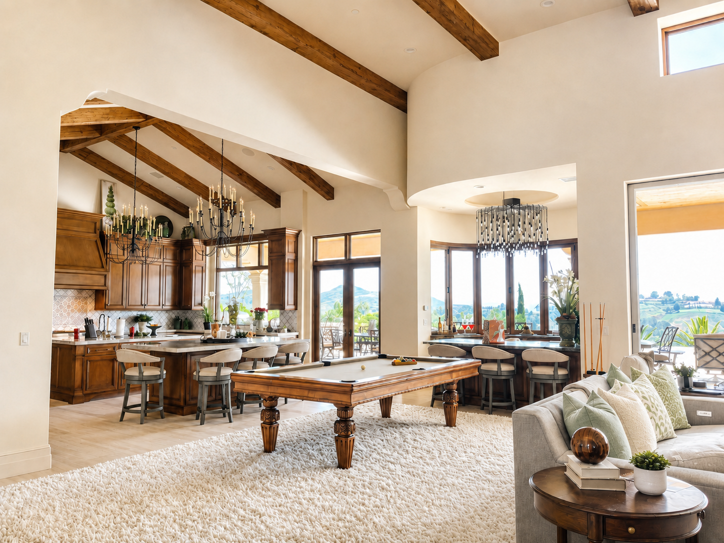Modern living room and kitchen with wooden ceiling beams, chandeliers, a pool table, and large windows showing a scenic outdoor view.