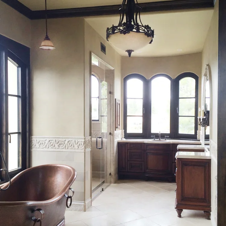 Bathroom with large windows, a copper clawfoot bathtub, a glass shower enclosure, and wooden cabinets. Natural light fills the space.