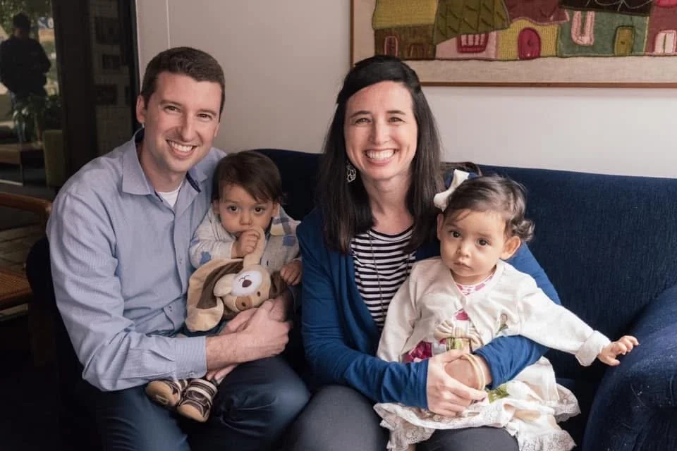 A happy family of four sitting on a couch, smiling at the camera. The father is on the left with a young girl sitting on his lap, holding a stuffed dog toy. The mother is on the right with her young daughter sitting on her lap. There is artwork on the wall behind them.