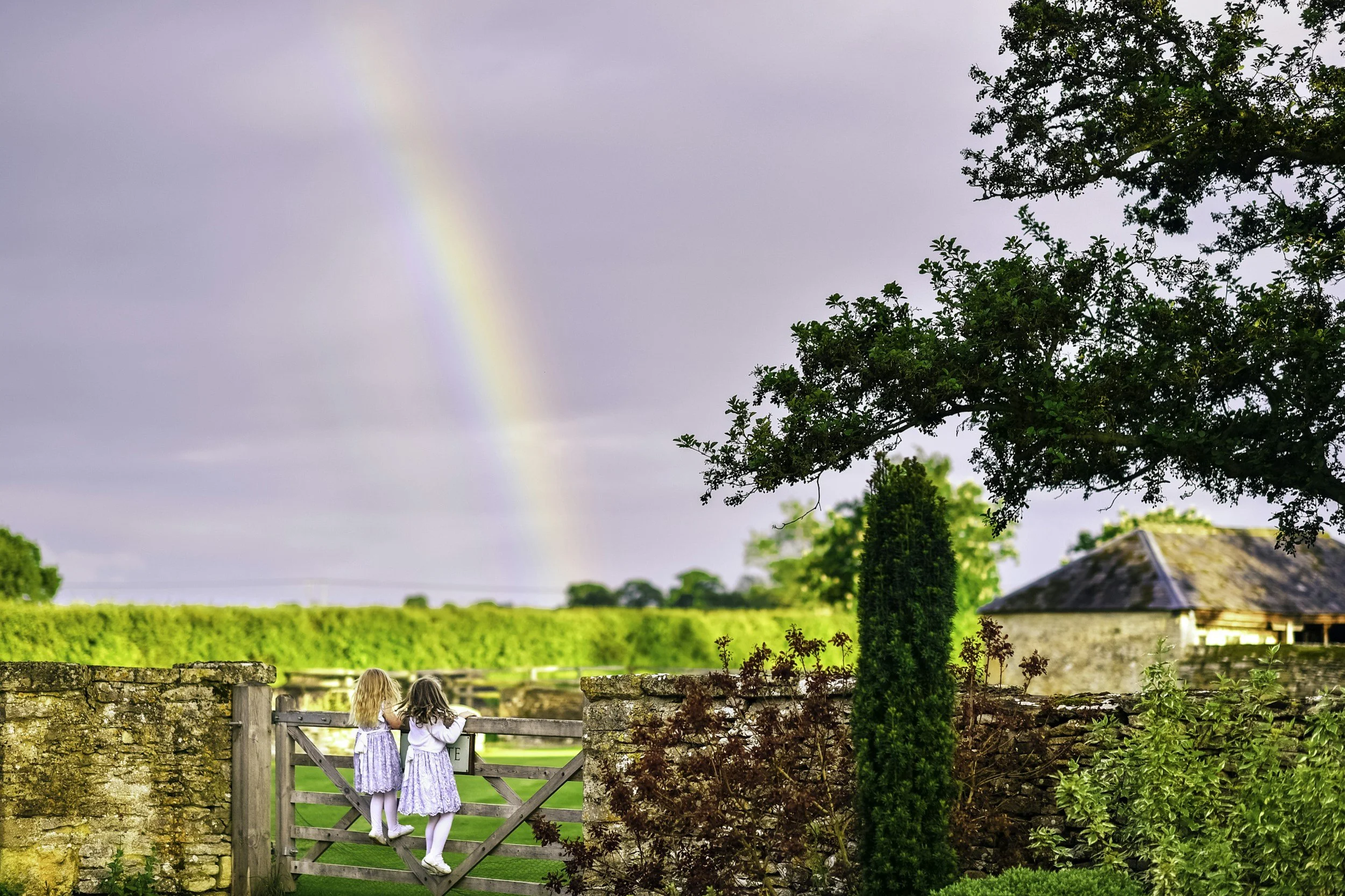 Two young girls in dresses standing by a wooden gate, looking at a lush green field, with trees, a house, and a rainbow in the sky.