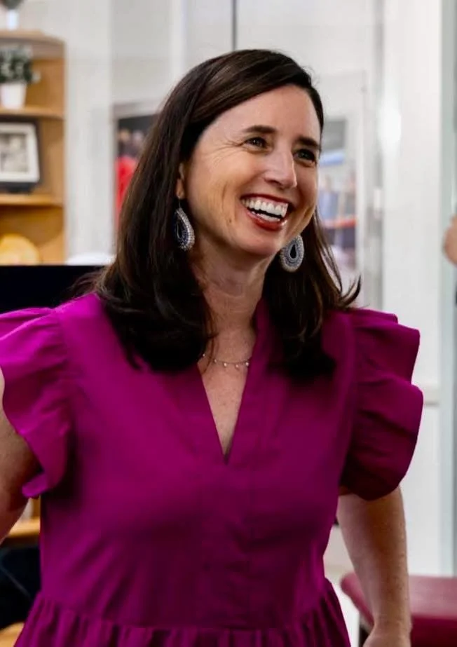 Dr. Johnson is shown with dark brown hair, wearing a magenta dress with puffed sleeves, smiling inside a bright room with bookshelves and framed photos in the background.