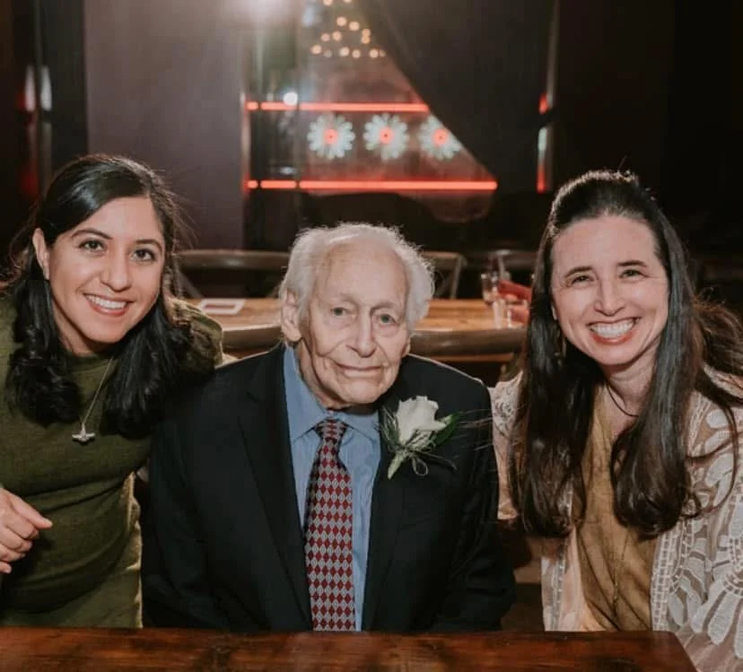 Dr. Richard Woodcock is pictured with Dr. Johnson a student sitting at a table in a restaurant or bar. The women are smiling. The elderly man has a neutral expression. The background has dim lighting and neon signs.