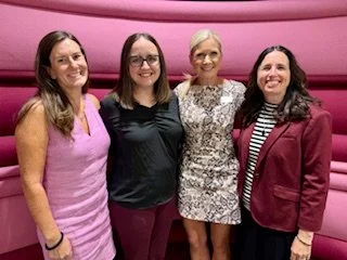 Four women standing together indoors in front of a pink upholstered wall, smiling for the photo.