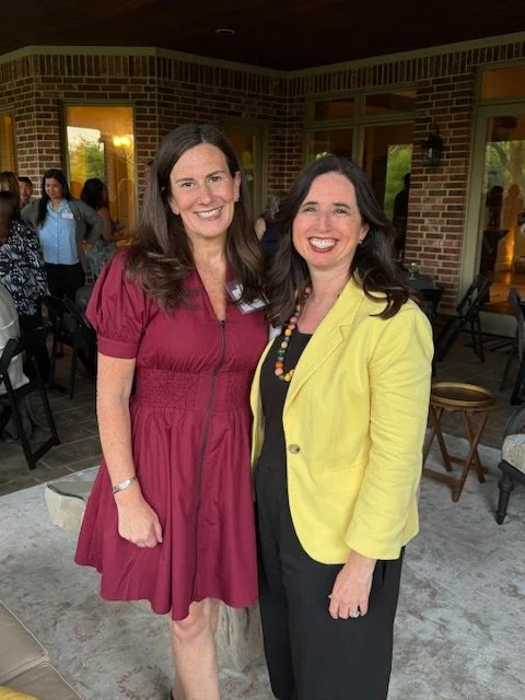 Two women standing together at an indoor gathering, smiling, with other people in the background. One woman wears a maroon dress, the other a yellow blazer over black clothing.