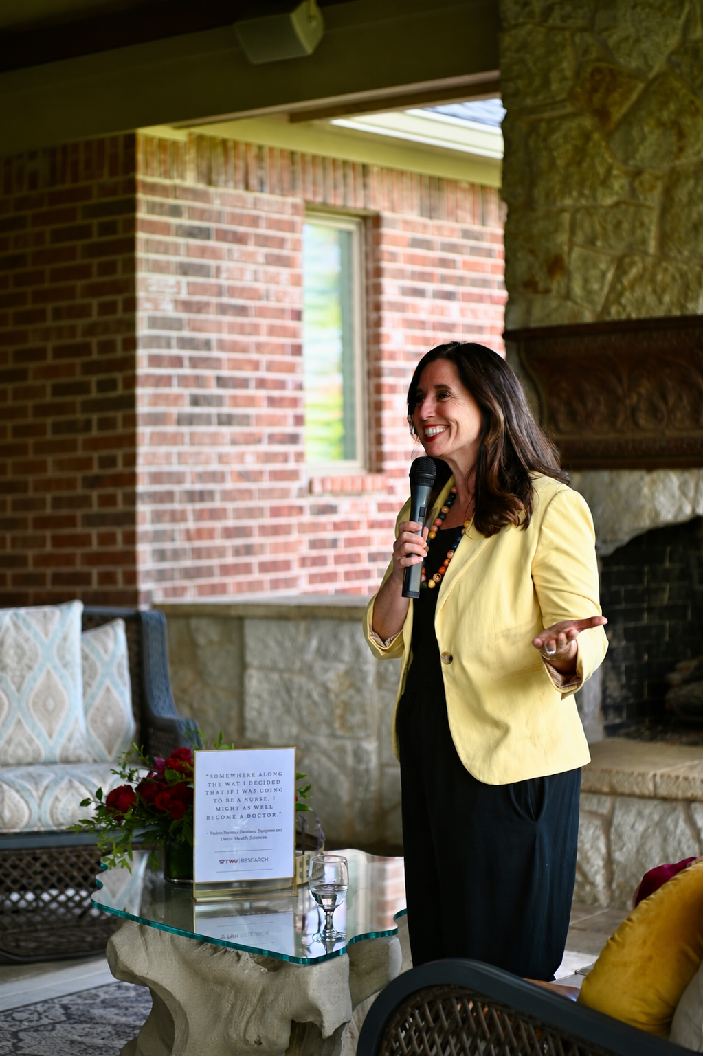 A woman with dark hair, wearing a yellow blazer and black dress, is speaking into a microphone in a cozy indoor setting with brick and stone walls, a fireplace, and wicker furniture. There is a small table with a glass top, a glass of water, a bouquet of red roses, and a sign.