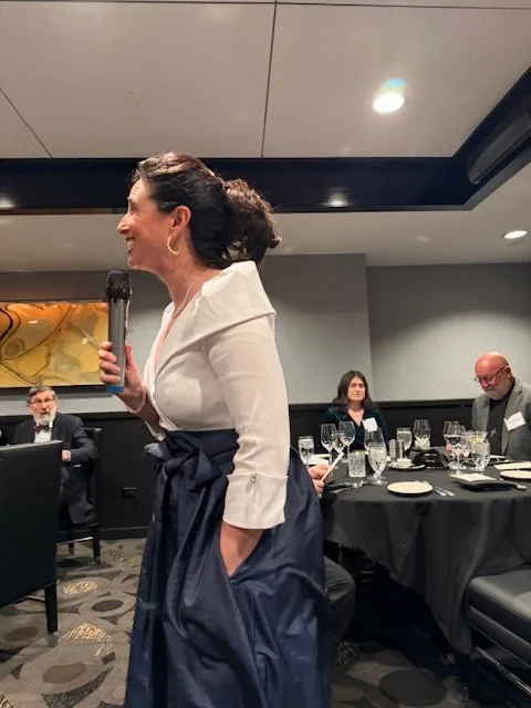 A woman standing and speaking into a microphone at a formal event, with three seated people at a table behind her in a conference room.