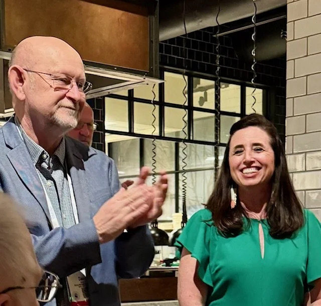 A man with glasses and a beard in a blue blazer clapping next to a woman with long dark hair in a green top, smiling in an indoor setting with a black and white tiled wall and large windows in the background.