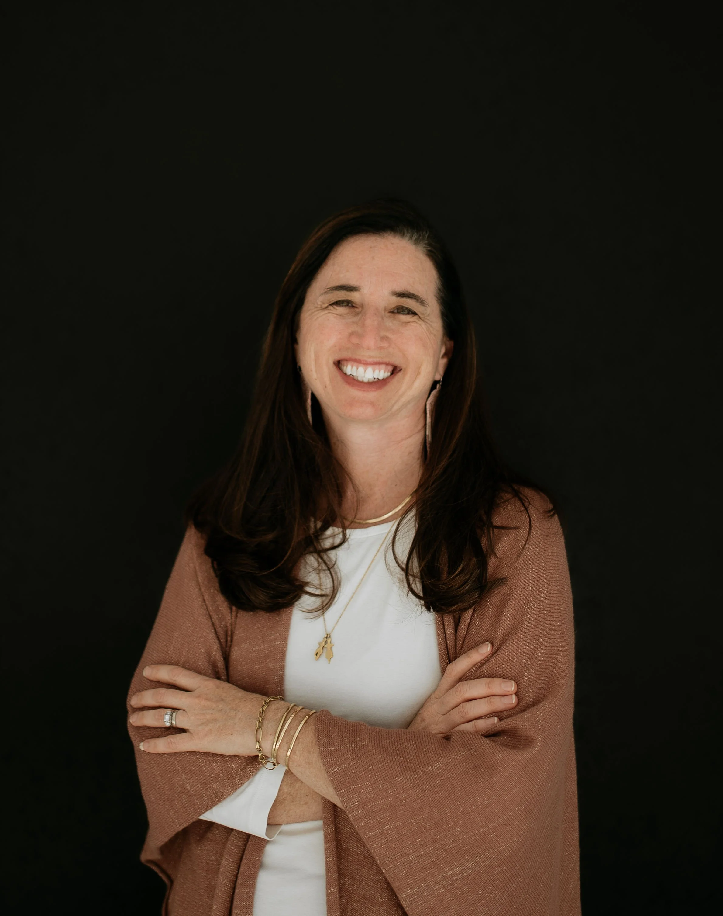 A woman with long brown hair smiling and crossing her arms, wearing a white top, a tan cardigan, and gold jewelry, against a black background.