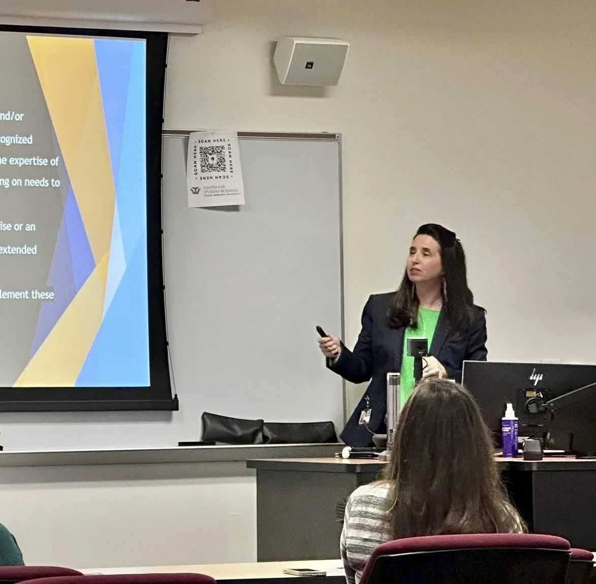 A woman giving a presentation in a classroom, standing next to a large screen displaying a colorful slide, with students seated facing her.