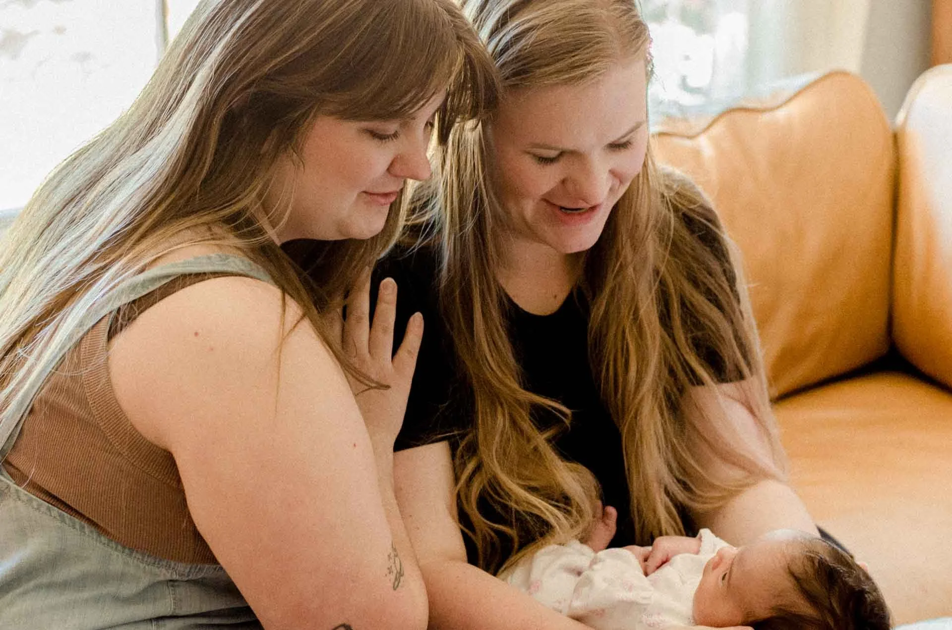 Two women with long red hair looking down at and smiling at a baby lying on a table. They are sitting on a brown bench with a light-colored wall and window in the background.