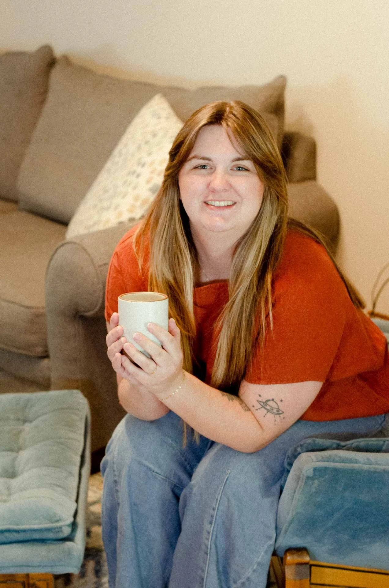 A smiling young woman with long, red hair and a tattoo on her right forearm, sitting on a couch holding a white mug with both hands, wearing a rust-colored top and blue jeans in a cozy living room.