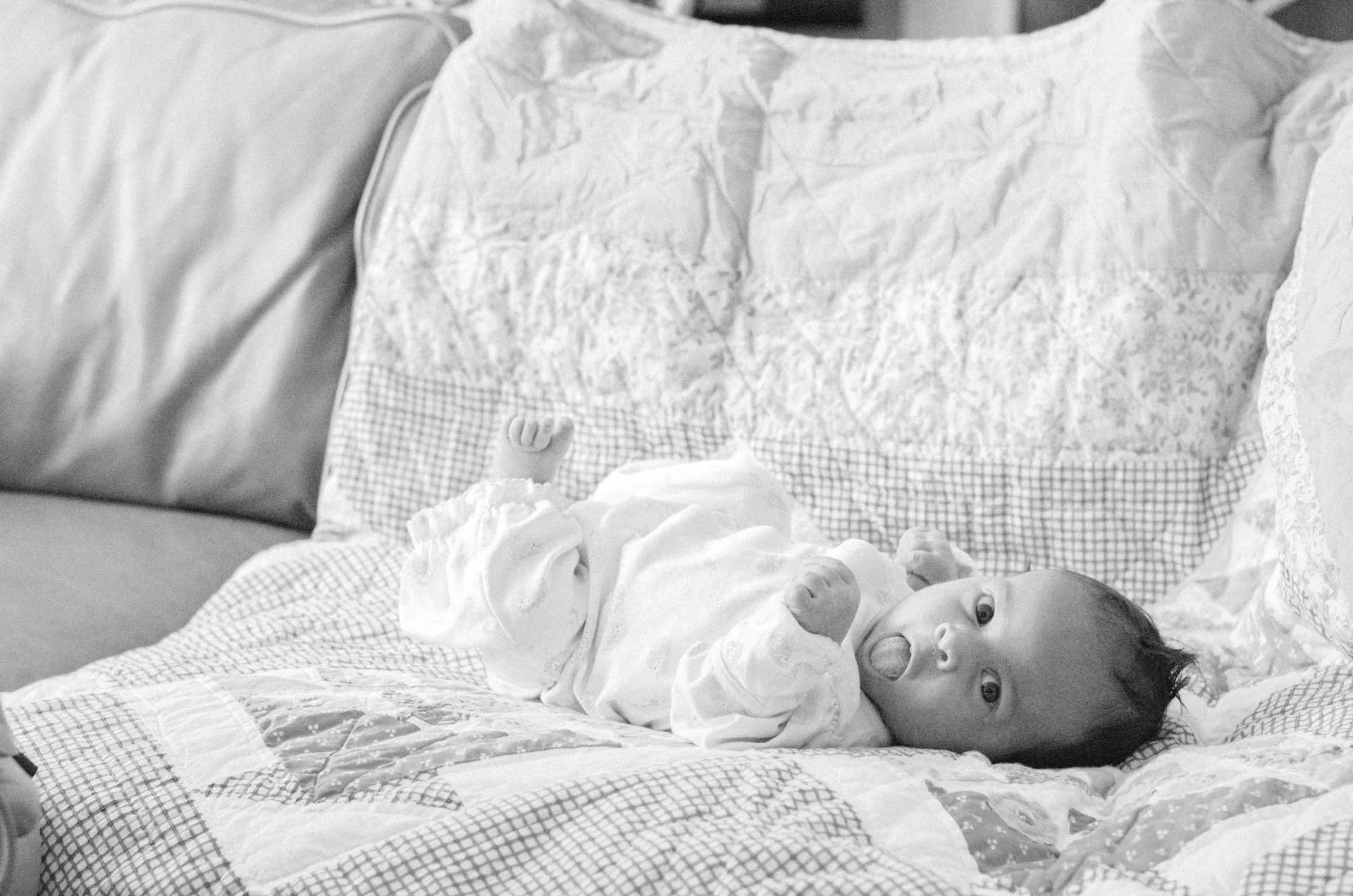 A baby lying on a hospital bed with is mouth open, wearing a white gown, with a pillow and blanket.