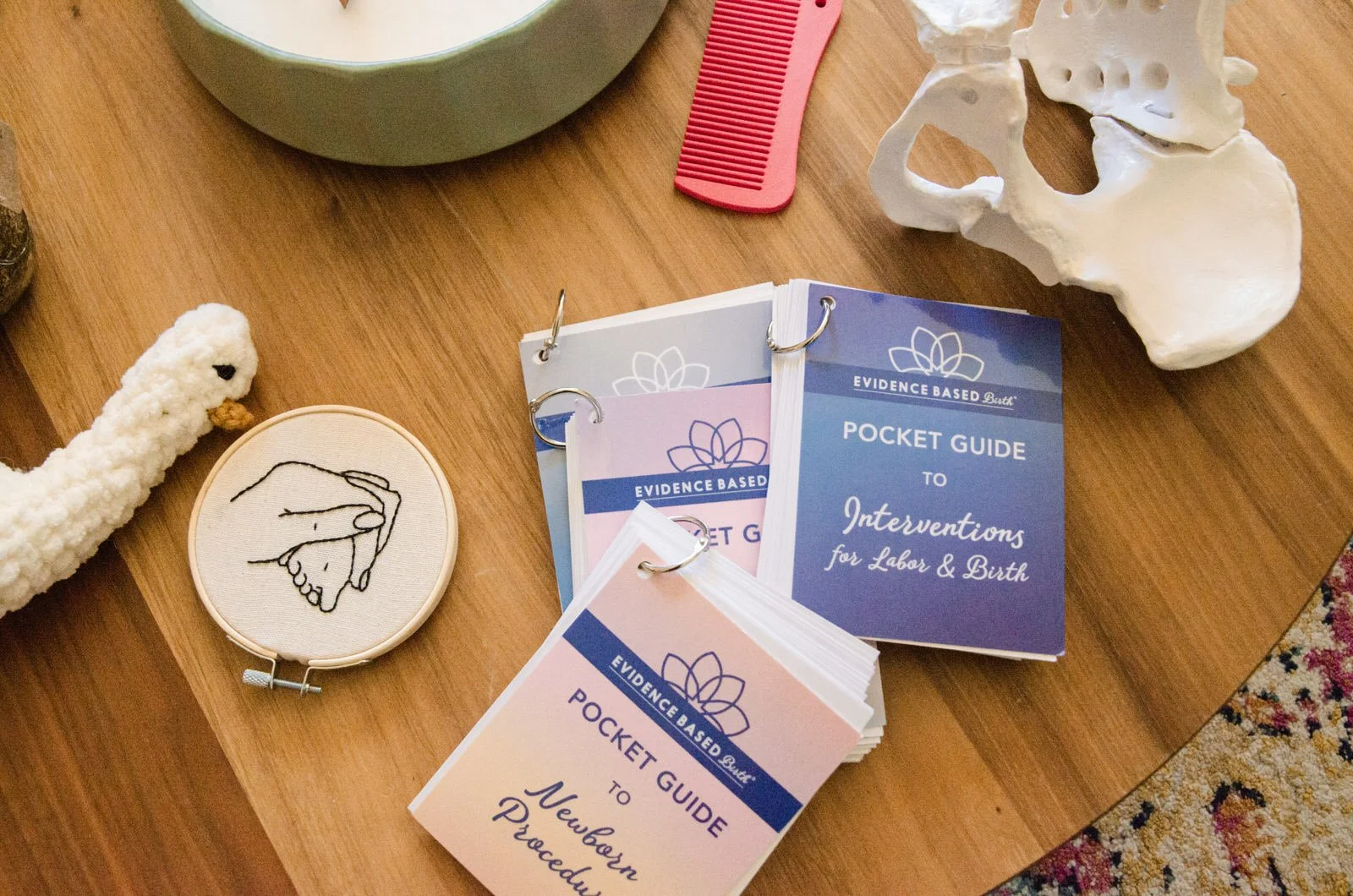 Table with medical and emotional aids, including pamphlets titled 'Pocket Guide to Interventions for Labor & Birth,' a white fabric chicken, a small embroidery hoop with a line drawing of two hands, a plastic pelvic skeleton model, a pink comb, and a green bowl.