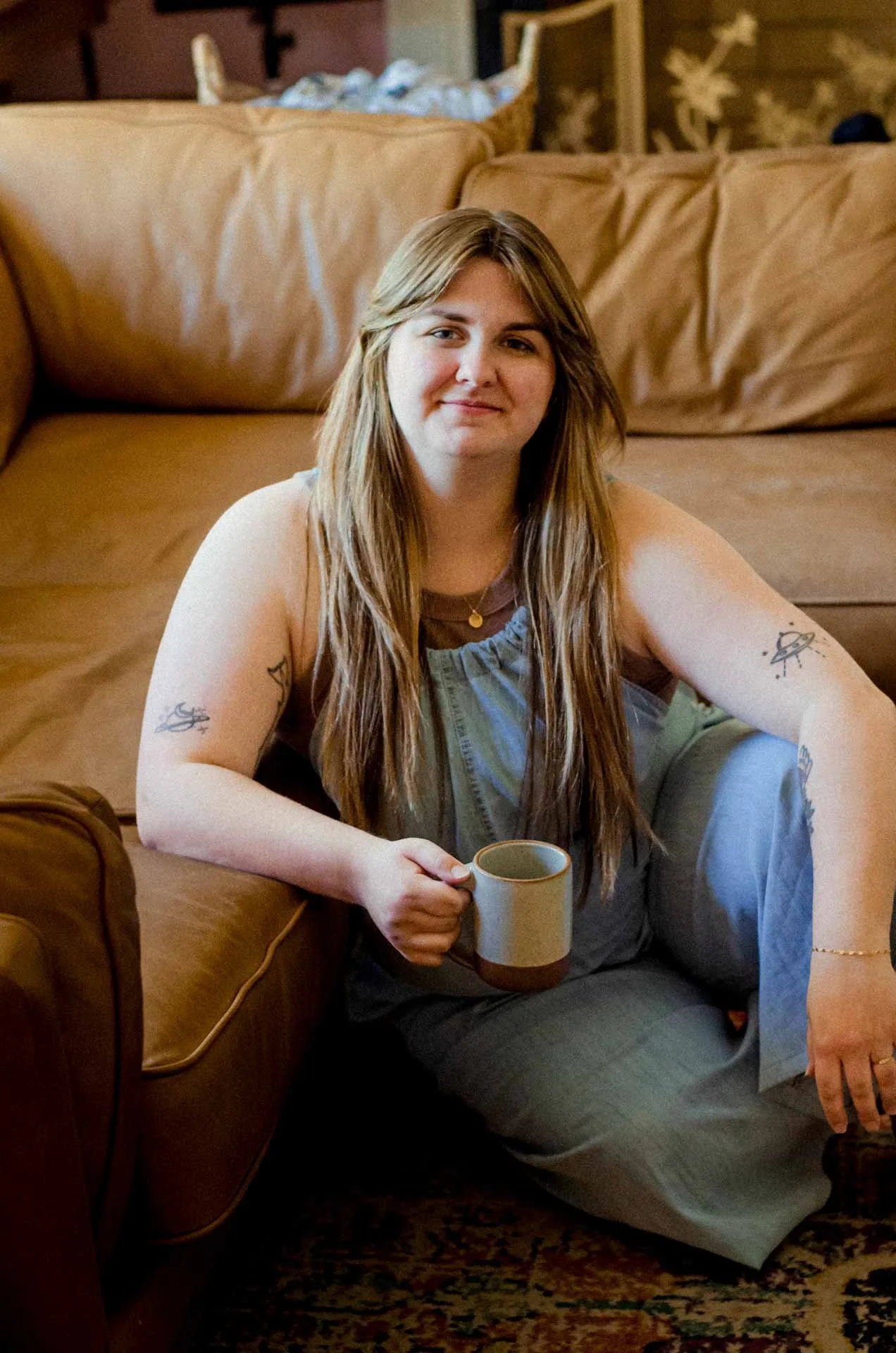 A young woman with long brown hair, tattoos on her arms, sitting on the floor beside a brown leather couch, holding a beige mug, smiling at the camera in a cozy living room.
