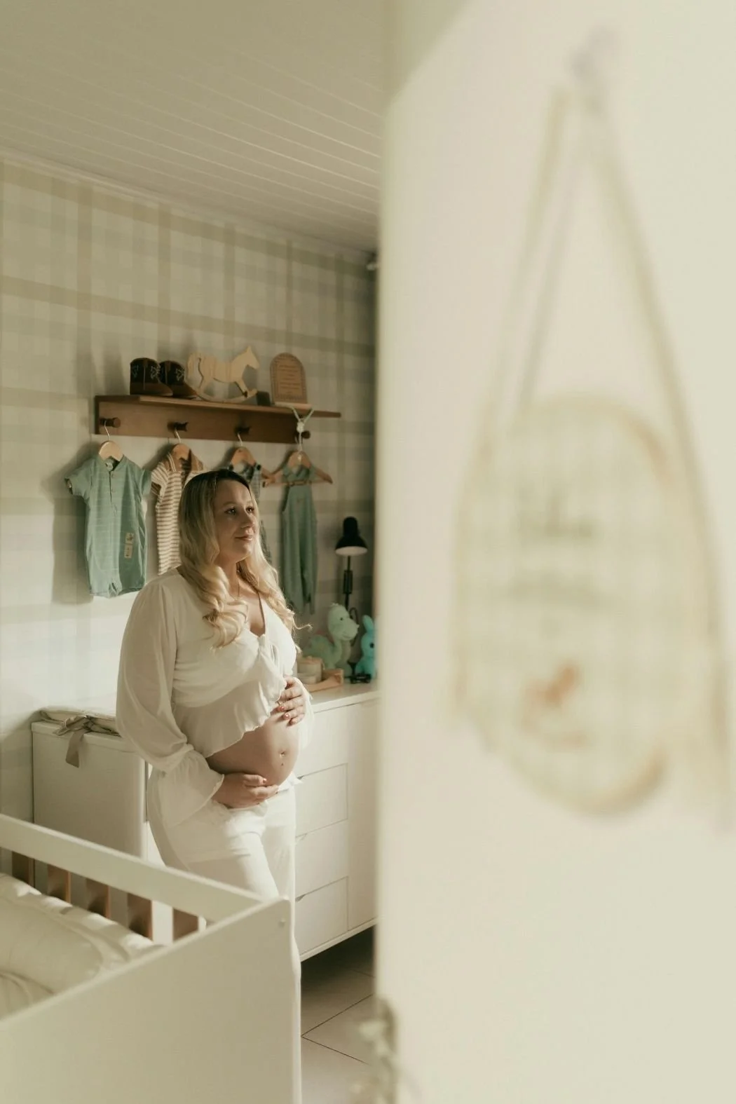 A pregnant woman standing in a nursery, gently holding her belly, with baby clothes hanging on a wall behind her.