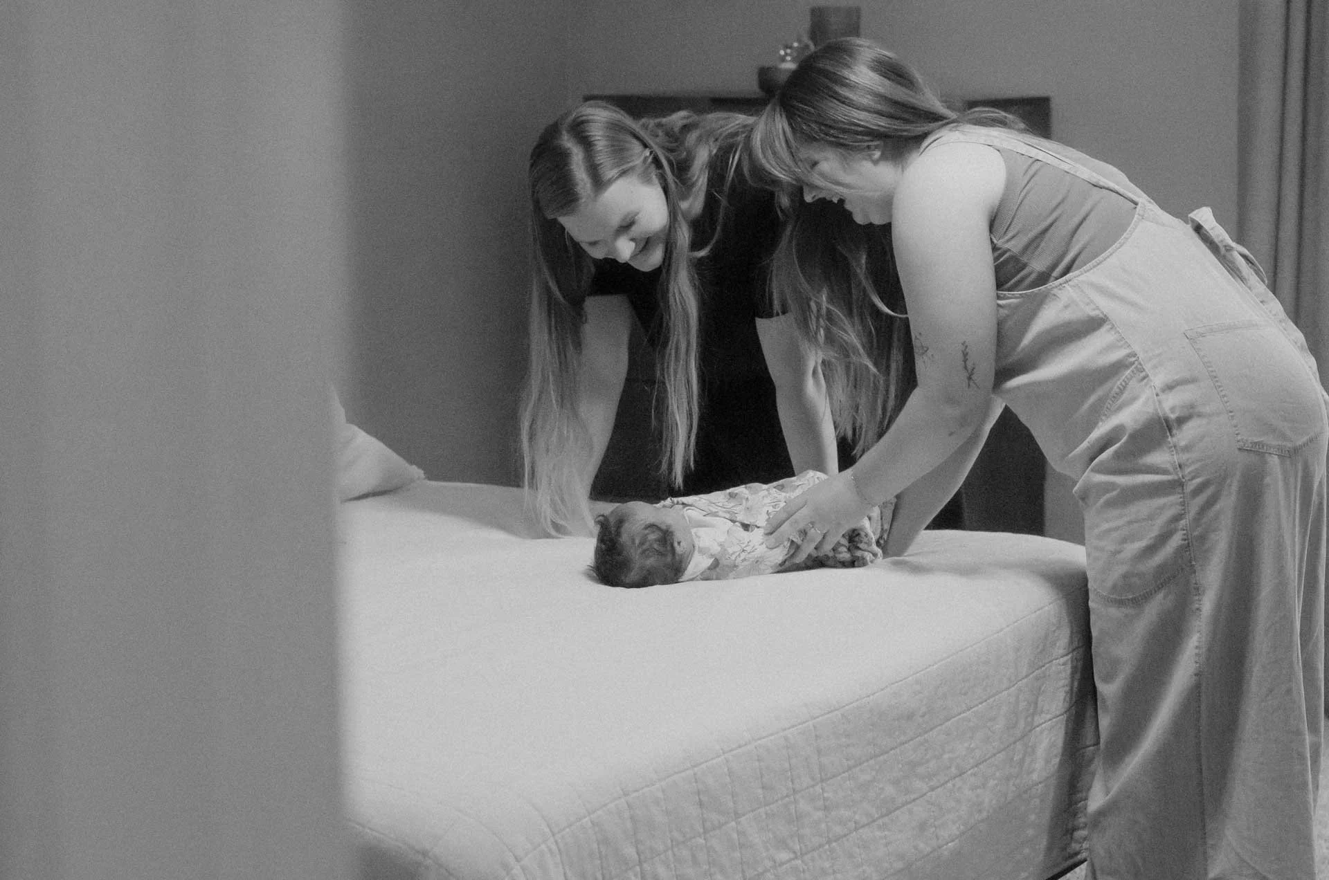 Two women are smiling and leaning over a bed, gently touching a small puppy wrapped in a floral blanket.