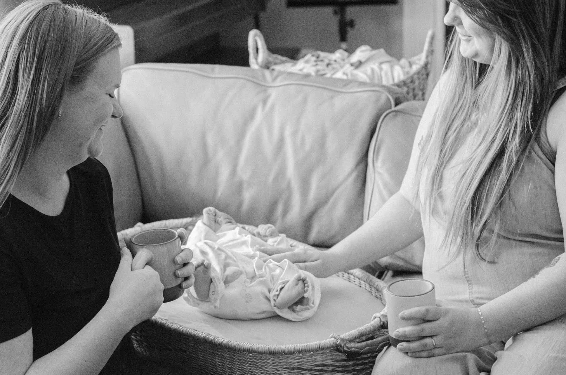 A woman with long hair in a sleeveless top is holding a mug and sitting beside a newborn baby wrapped in a blanket. Another woman with long hair, wearing a bracelet and a sleeveless top, is gently holding the baby's hand. The scene is in a cozy room, possibly a nursery, with a bed or changing table in the background.