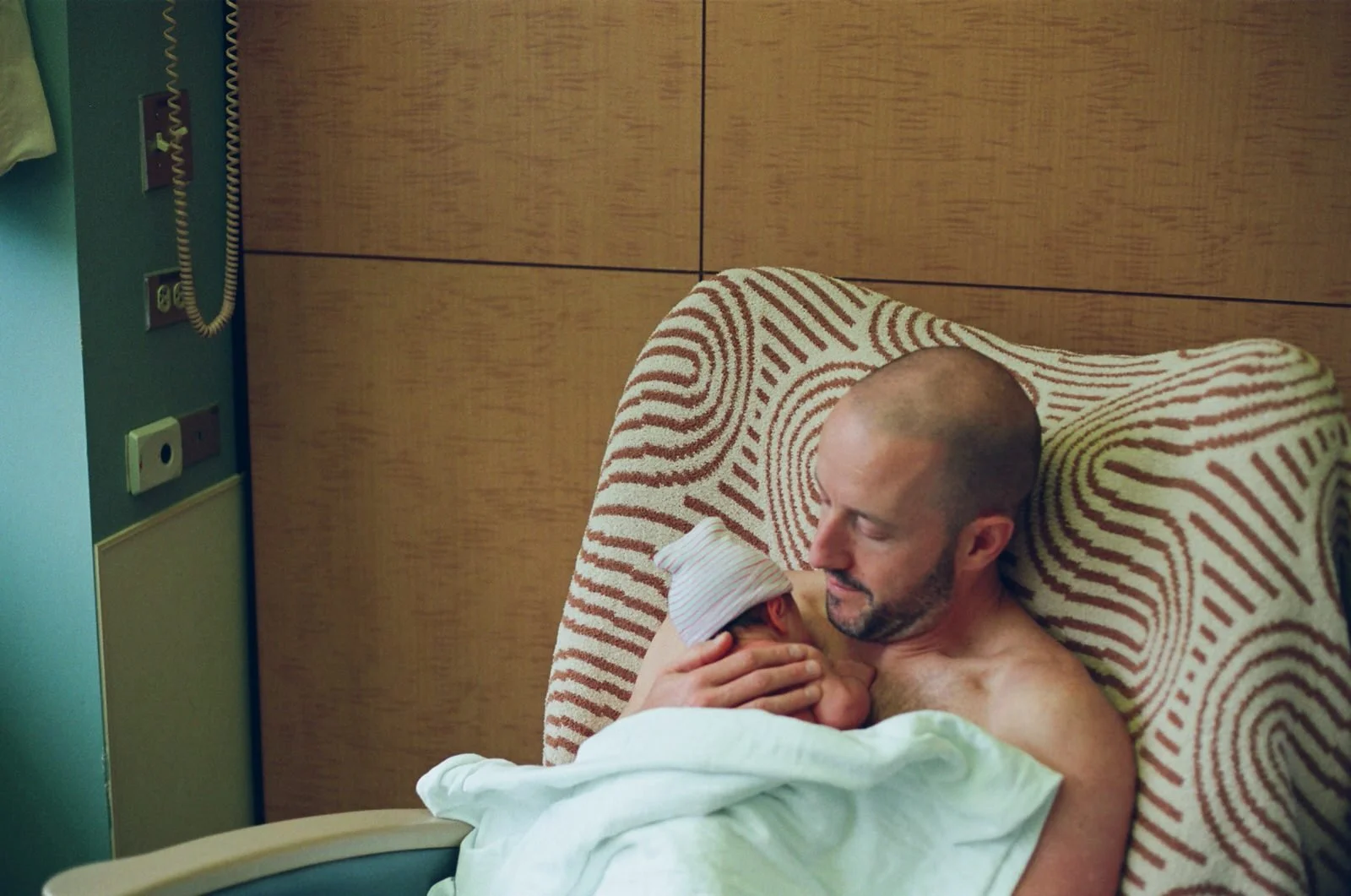 A man holding a newborn baby while sitting in a hospital bed with a patterned headboard. The man is smiling and looking at the baby, which is wearing a striped hat.