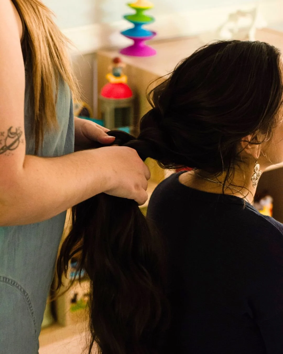 A woman with long dark hair is having her hair styled or braided by another person with light hair and a tattoo on her arm, in a room decorated with colorful toys and objects.