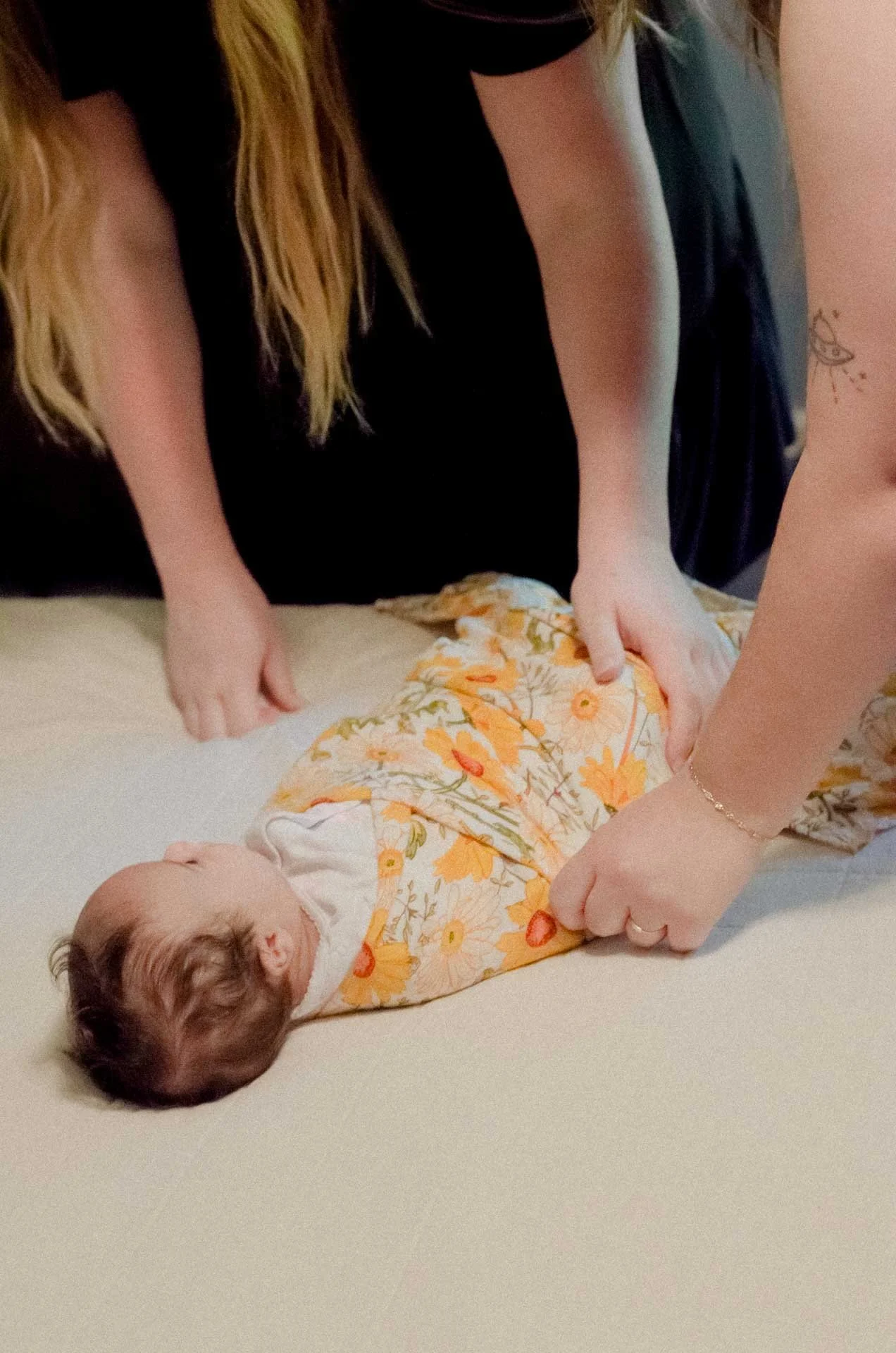A person lying on their back with a baby on top, wearing a floral onesie and surrounded by two adults who are holding and supporting the baby.