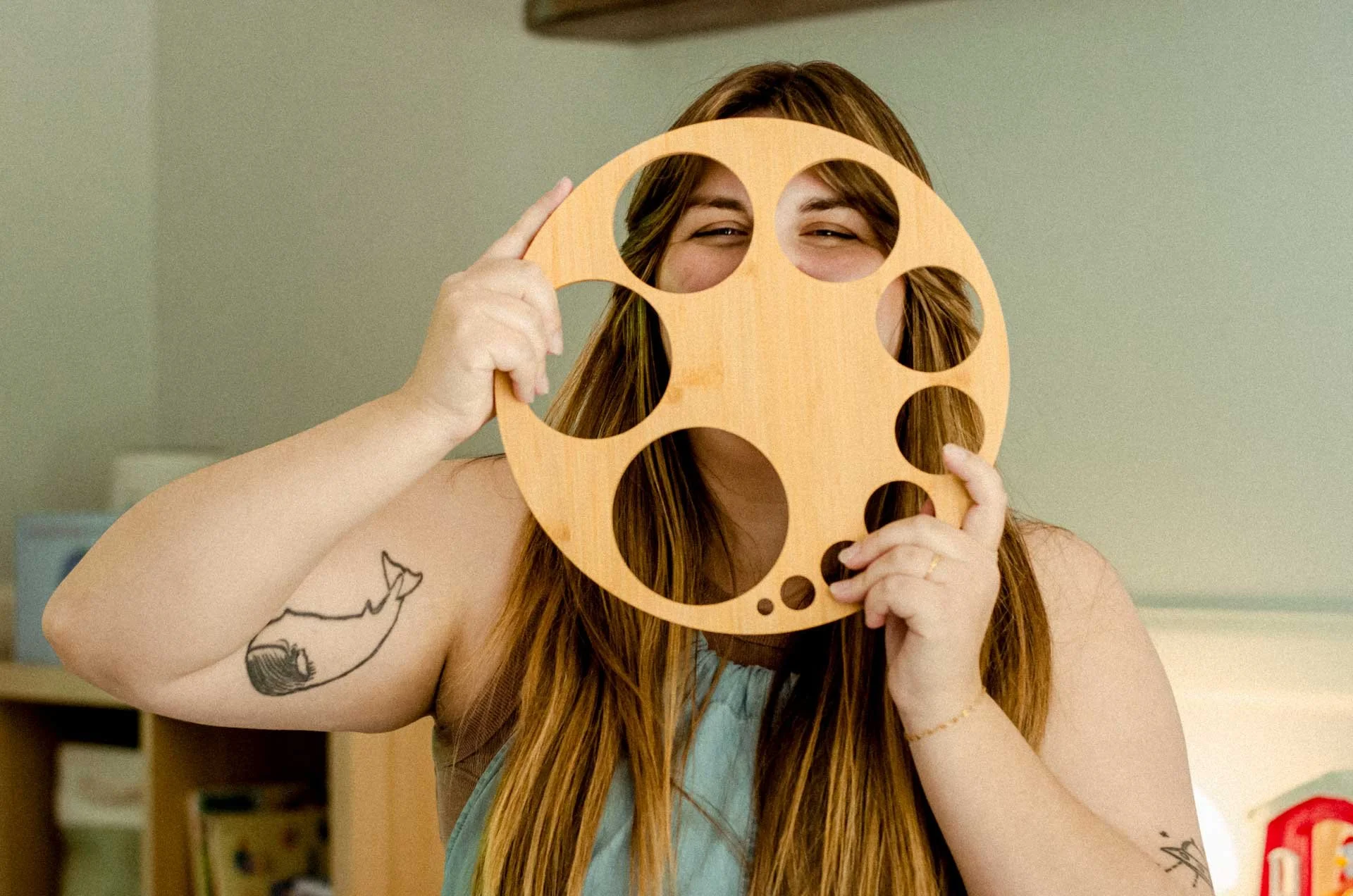 A woman with long hair holding a wooden circle with multiple holes in front of her face, smiling. She has tattoos on her arm and a bracelet on her wrist.