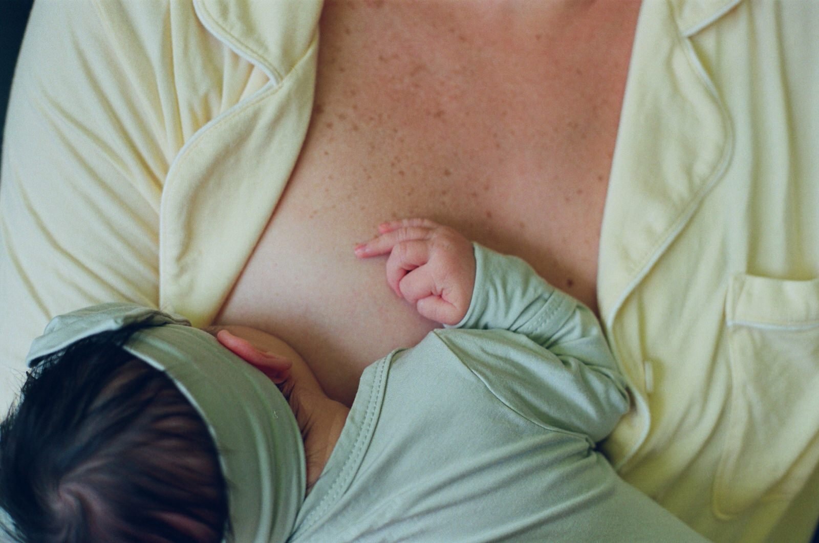 A woman in a pale yellow robe breastfeeding a baby who is wearing a light green shirt, lying on her chest.