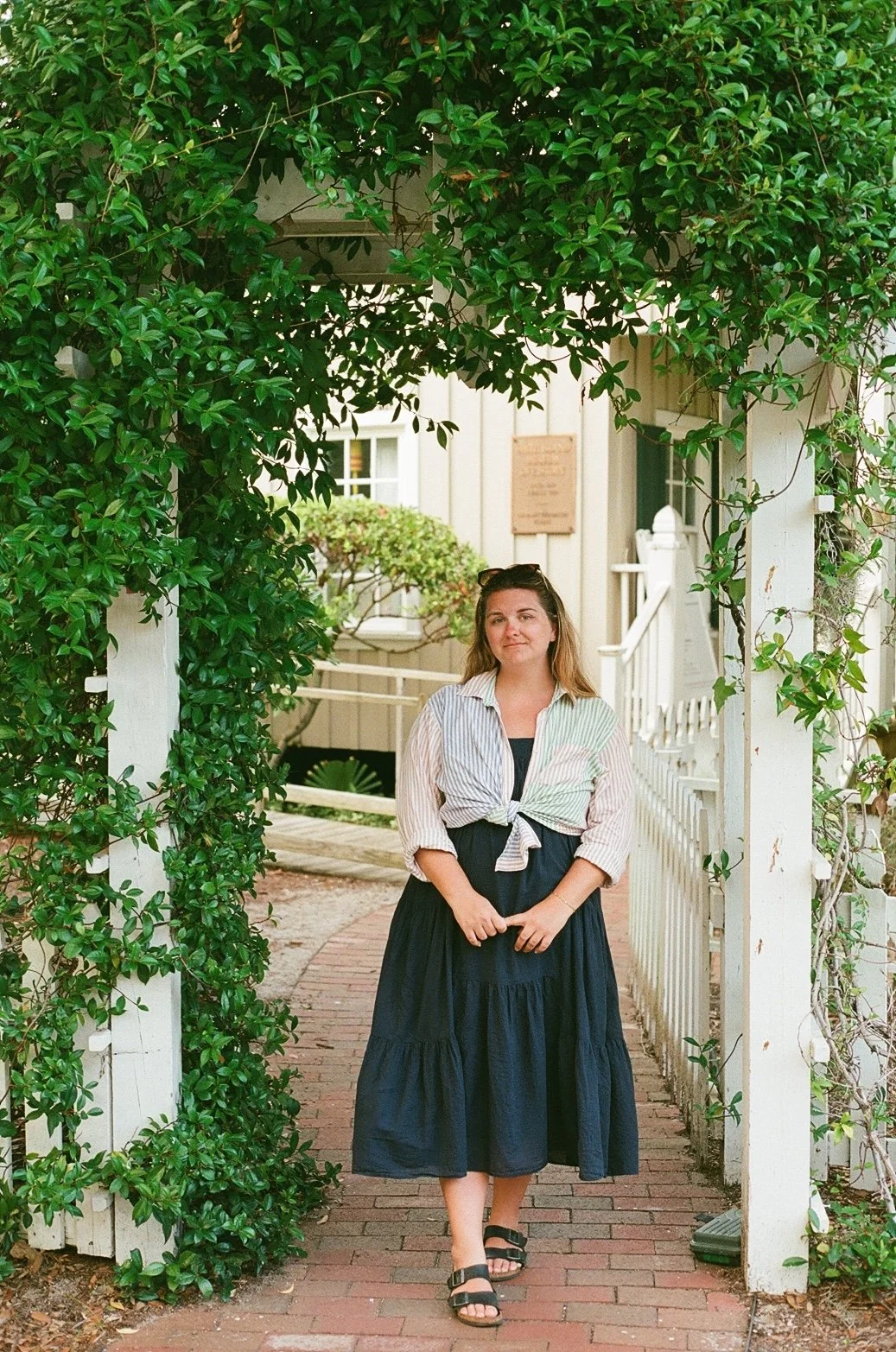 A woman standing under a leafy archway at a historical site, wearing a layered skirt and a knotted shirt.