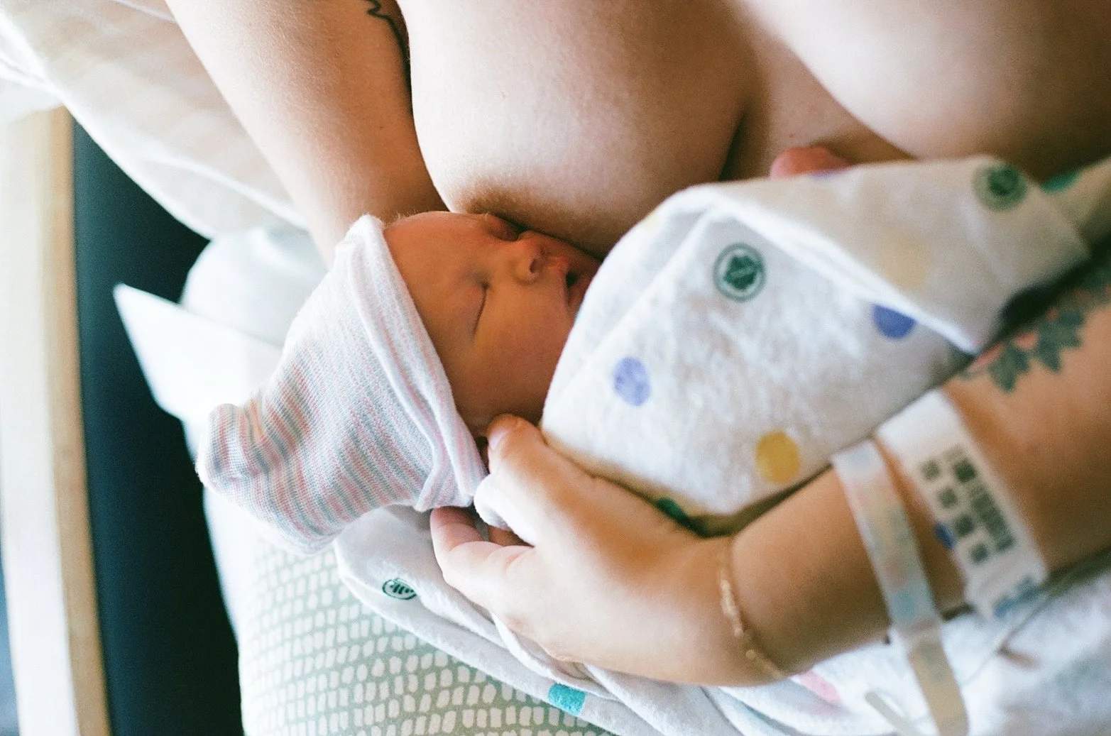 Newborn baby breastfeeding while wearing a pink and white striped hat, held by a person with a hospital wristband, lying in a hospital bed.