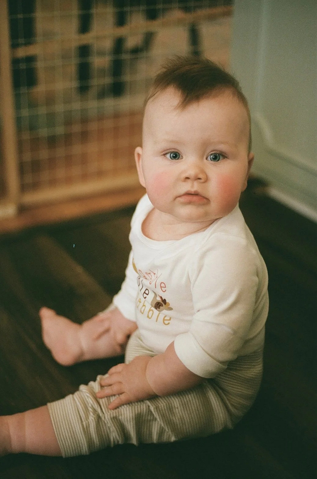 A young child with light skin, chubby cheeks, and brown hair spiked at the top, sitting indoors on a dark surface, wearing a white shirt with colorful text and beige striped pants, with a neutral expression, looking up at the camera.